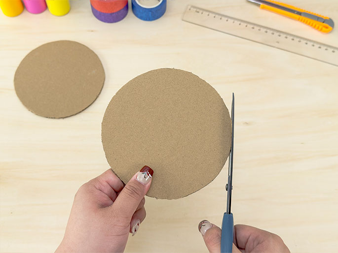 Two hands are shown manipulating circular pieces of cardboard on a light woodgrain surface. One hand holds a cardboard circle while the other hand uses scissors to trim the edge of the circle. Another identical cardboard circle lies fully cut on the table nearby. This scene suggests a crafting activity involving cutting and preparing cardboard shapes, with other crafting materials like colorful tape, a ruler, and a utility knife visible in the background. The fingernails of the hands holding the scissors and cardboard are painted with a decorative design.