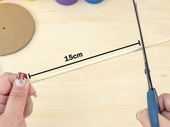 Two hands are in the process of cutting a thin wooden dowel with scissors. The dowel is measured at 15cm, indicated by a black line and text label. The hands hold the dowel and scissors against a light woodgrain background. A partially completed cardboard circle is visible in the top left corner, suggesting this action is part of a crafting sequence. The fingernails of the hands holding the scissors and dowel are painted with a decorative design.