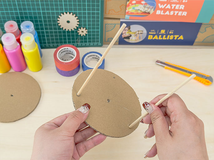Two hands are assembling a craft project involving a circular piece of cardboard with two wooden dowels inserted through small holes near its edge. A second, identical cardboard circle lies on the light woodgrain work surface to the left. Various crafting materials, including bottles of paint, rolls of colorful tape, wooden gears, a utility knife, and boxes labeled WATER BLASTER and BALLISTA, are visible in the background. The fingernails of the hands holding the dowels and cardboard are painted with a decorative design.