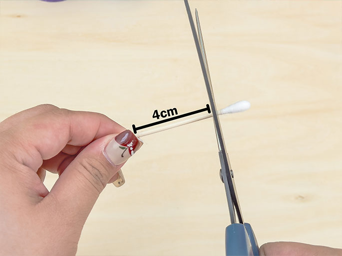 Two hands are shown cutting a cotton swab with scissors. The swab's stick, measured at 4cm and indicated by a black line and text label, is being trimmed on a light woodgrain surface. The hand holding the cotton swab has fingernails decorated with a floral design. The focus on the cutting action suggests this is a step in a crafting or DIY process.