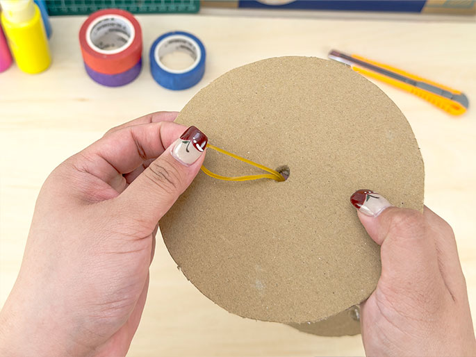 Two hands hold two stacked circular pieces of cardboard. A yellow rubber band is looped through a small hole in the center of the top cardboard circle. The hands, with fingernails decorated with a floral design, manipulate the rubber band. A utility knife, rolls of colorful tape, and bottles of paint are visible in the background on the light wooden work surface, suggesting this is a step in a crafting project.