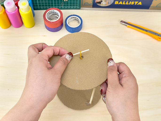 Two hands are working on a craft project. They are placing a short piece of a cotton swab stick through a hole in the center of a circular piece of cardboard. A yellow rubber band is also looped through the same hole. The cardboard circle rests on a cylindrical cardboard structure supported by wooden dowels. The background includes craft supplies like paint, tape, and a utility knife on a light wood surface. The fingernails of the hands working on the project are painted with a decorative design. A box labeled BALLISTA is also visible in the background.