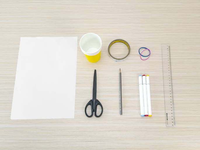A flatlay image displays an assortment of craft or school supplies arranged on a light wood-grained table. The items include a blank sheet of white paper, a small yellow disposable cup, a roll of clear tape, a pair of linked rubber bands, a pair of black scissors, a pencil, three white markers with colored caps (light blue, red, and yellow), and a clear plastic ruler. The scene suggests preparation for a creative project or activity.