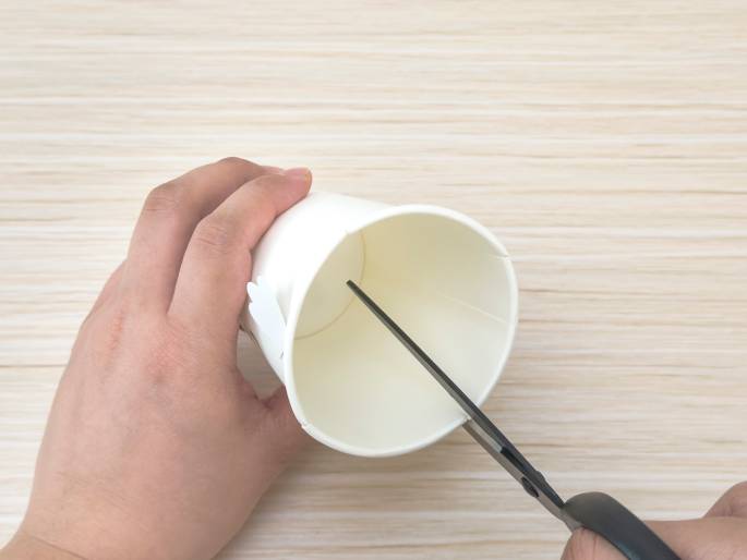 Two hands are manipulating a white paper cup on a light wood-grained surface. One hand holds the cup, which is viewed from above, revealing its interior. The other hand holds a pair of scissors and is in the process of making a cut inside the cup. Small, white, wing-like decorations are visible on the side of the cup. This suggests a craft activity where the cup is being modified or prepared for a specific purpose.