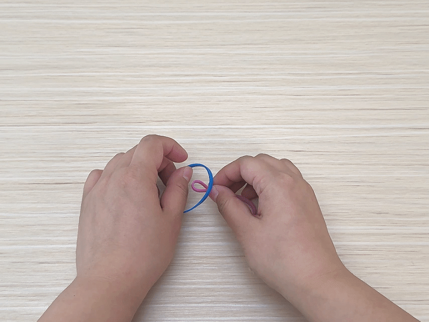 Against a light wood-grained background, two hands are shown manipulating a small blue rubber band and a smaller pink rubber band. The blue rubber band is larger and forms a loop. The pink rubber band is threaded through the blue one, creating a small linked section. The hands appear to be in the process of connecting or weaving the rubber bands together, likely as part of a small craft or activity like making a bracelet or other item from rubber bands.