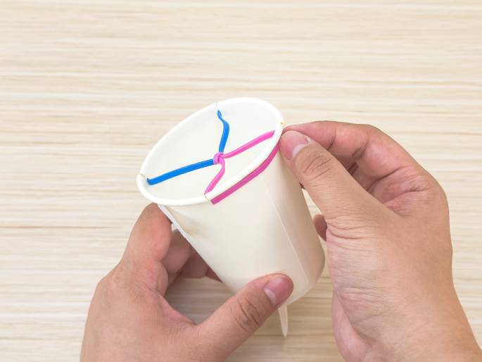 Two hands hold a white paper cup against a light wood-grained background. Pink and blue rubber bands are stretched across the opening of the cup, intersecting in the center. The rubber bands appear to be holding something in place or creating a type of launcher or catapult mechanism. Small cuts or slits are visible in the rim of the cup, likely made to accommodate the rubber bands. A small, pointed object protrudes from the bottom of the cup, possibly a piece of plastic or wood, adding to the impression that this is a homemade device or toy.