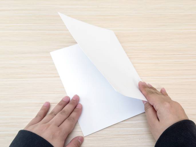 Two hands, partially covered by dark sleeves, hold a sheet of white paper that is being folded in half. The paper rests on a light wood-grained surface. The image captures the moment of the fold, with the top portion of the paper lifted and about to be creased against the bottom portion. This simple action suggests the beginning of a paper craft, origami, or letter-writing activity.