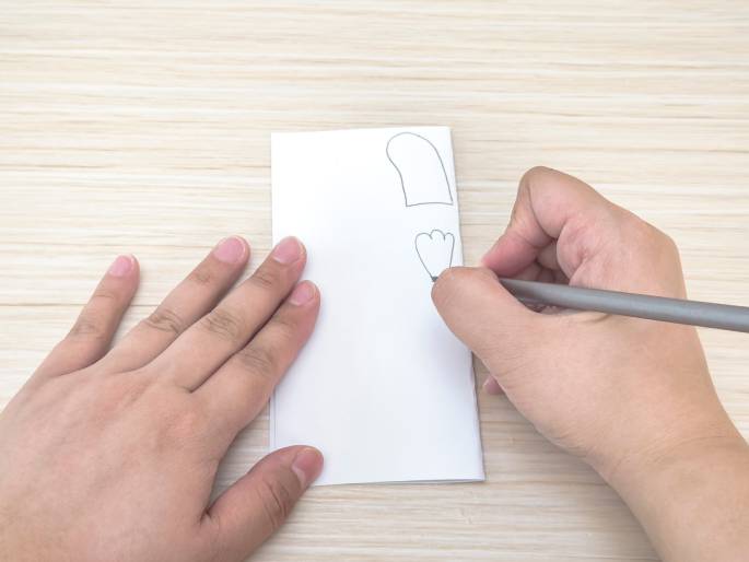 On a light wood-grained surface, a piece of folded white paper lies flat. One hand holds the paper steady while the other hand, holding a pencil, draws small, simple shapes on it. Two shapes are already visible: one resembles a rounded rectangle or a mitten, and the other looks like a three-petal flower or a simplified paw print. The image suggests the initial steps of drawing or designing something, possibly a craft project.