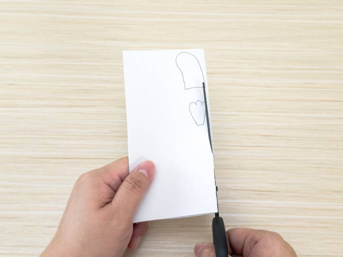 Two hands are engaged in a paper-cutting activity on a light wood-grained table. One hand holds a folded piece of white paper on which a mitten-like shape and a three-petal flower or paw print shape are lightly drawn. The other hand holds a pair of scissors and is in the process of cutting along the edge of the paper. The image captures a moment of crafting or creating a paper cutout.