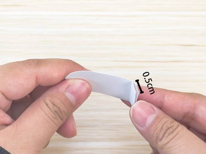 Two hands delicately fold a small strip of white paper. The paper, with a slightly rounded edge on one side, is being folded precisely. A black bracket and the text 0.5cm indicate a small, specific measurement for the fold, emphasizing the precision required for this step, likely part of a paper craft or origami project. The background is a light wood-grained surface.
