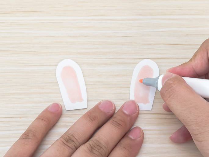 On a light wooden surface, two elongated, rounded paper cutouts, resembling animal ears, are being colored. One hand holds a light-colored marker with an orange tip and is applying a light pink or peach color to the inner part of one cutout. The other hand is positioned near the other cutout, which has already been colored similarly. The image suggests a crafting activity, possibly making animal ears for a costume or decoration.