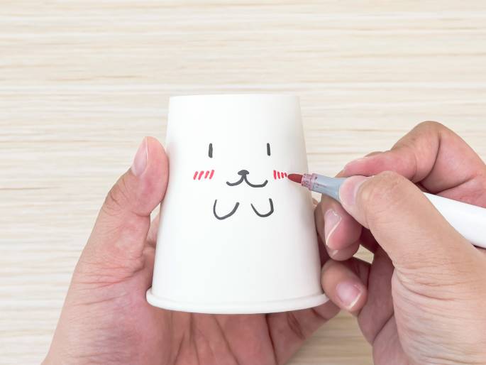 Against a light wood-grained background, two hands hold a white paper cup. A simple, cartoonish animal face is being drawn on the cup. One hand holds the cup while the other hand uses a marker to add red details, likely rosy cheeks, to the face. The drawing consists of short vertical lines for eyes, a small triangle for a nose, a curved line for a mouth, and three parallel red lines on each side for the cheeks. The image captures the process of decorating or personalizing a simple paper cup.