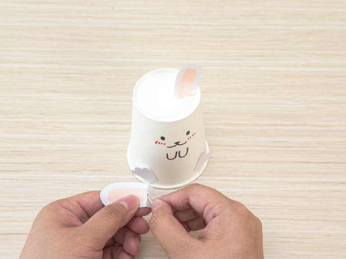 On a light-colored wooden table sits a white paper cup decorated with a cute animal face, featuring simple eyes, nose, mouth, and rosy cheeks. One ear-shaped cutout is already attached to the cup. Two hands are in the foreground, preparing to attach the second ear cutout, which has a small folded piece at its base, presumably for gluing or taping. This image shows the final stages of a simple craft project, creating an animal-themed cup.