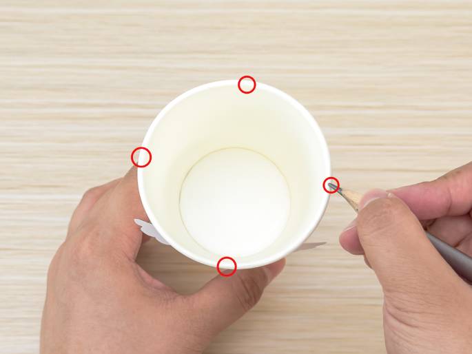 Two hands interact with a white paper cup on a light wood-grained surface. The cup is viewed from above, showing its interior. One hand holds the cup while the other hand uses a pencil to poke a small hole near the rim. Four red circles highlight locations around the rim of the cup, indicating designated spots for creating holes, likely for a crafting project. Small, white, wing-like shapes are visible on the sides of the cup, suggesting it's being transformed into an animal or creature.