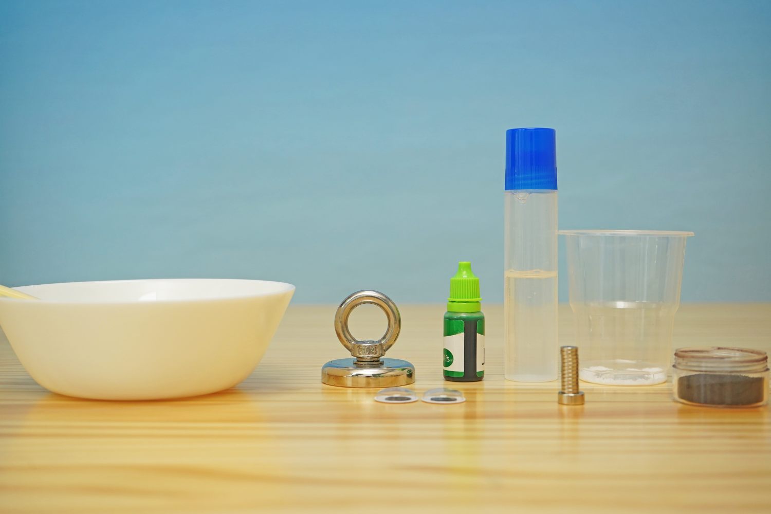 Gather your ingredients for some seriously gooey fun: a bowl, spoon, strong magnet with ring, green food colouring, PVA glue, water, small screw, and magnetic powder. Let's get started!