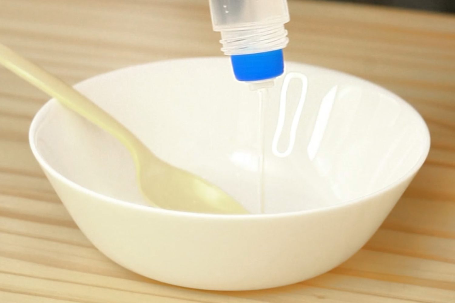 A hand pours clear glue from a bottle into a white bowl, a pale yellow spoon rests inside, all on a light wood surface. Let's get that slime started!