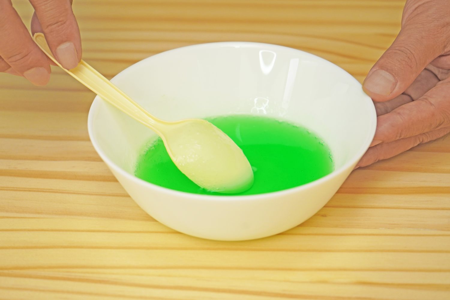 A pale yellow spoon rests in a bowl of bright green liquid, held by a hand on a light wood surface. Time to get mixing!