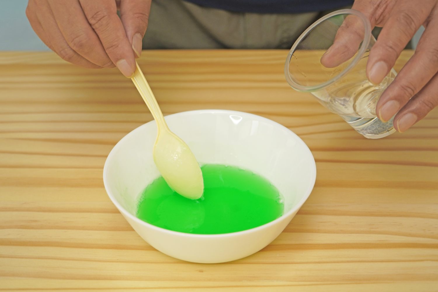 A hand pours water into a bowl of green liquid while another hand stirs with a spoon, all on a light wood surface. The mixture is getting ready for the next stage of slime-making.