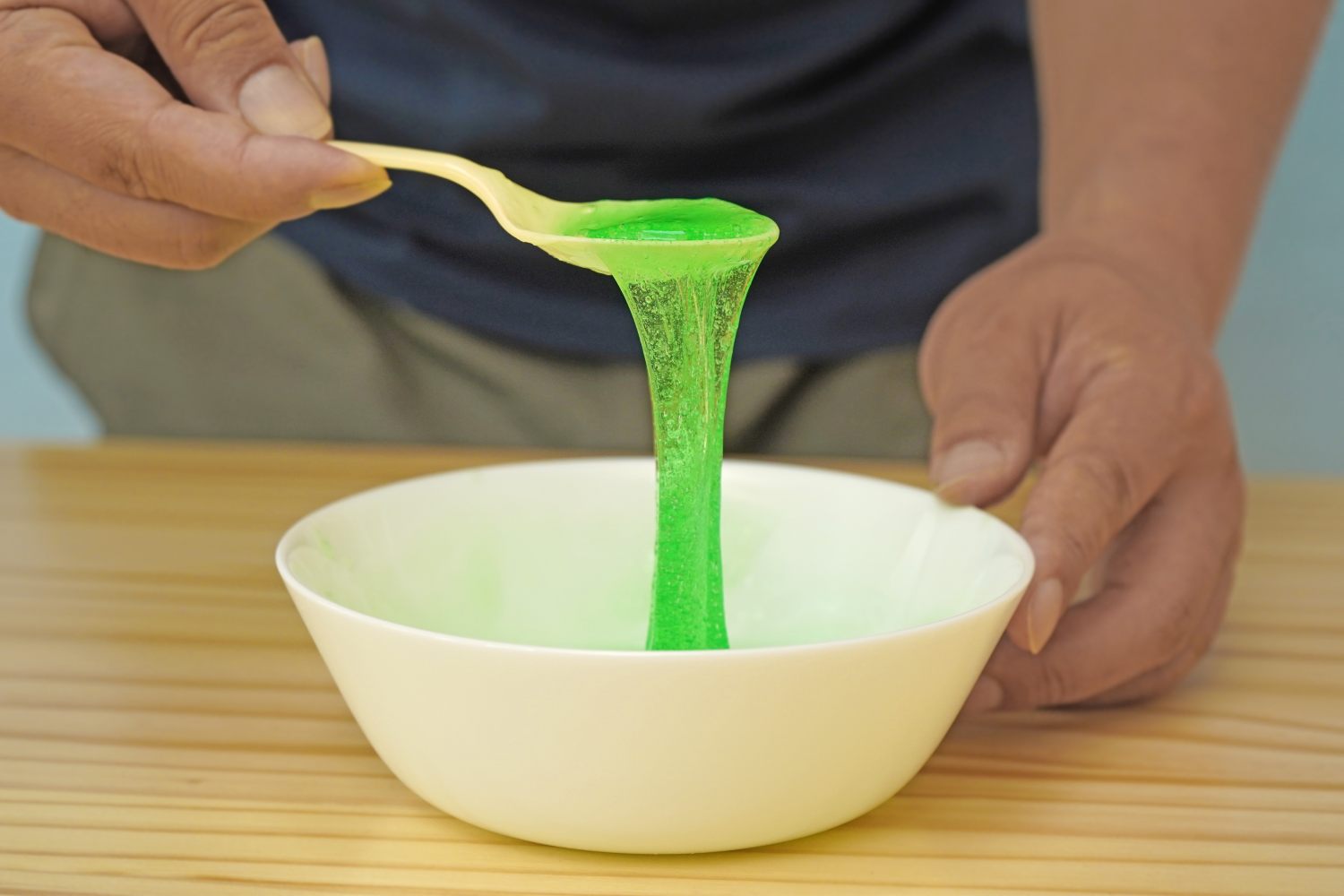 Ooh, look at that satisfying slime! A person is carefully pouring bright green slime from a spoon into a white bowl on a light wood surface. Nearly ready for the magnetic fun!