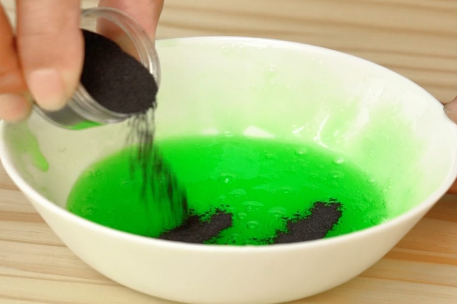 Adding the secret ingredient! A hand pours dark grey magnetic powder from a small container into a bowl of bright green slime, on a light wood surface. Nearly there!