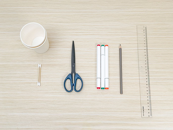 An assortment of craft supplies is neatly arranged on a light wooden table. The items include a small paper cup, two cotton swabs, a pair of scissors, three white markers with green, orange, and red caps, a pencil, and a clear plastic ruler. The overhead view suggests these items are ready to be used for a craft project.