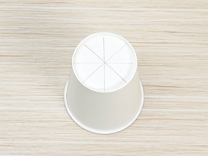 A small white paper cup is placed upside down on a light wooden surface. The bottom of the cup has lines drawn on it, dividing it into eight equal sections, like slices of a pie. This suggests preparation for a craft project involving cutting or folding.