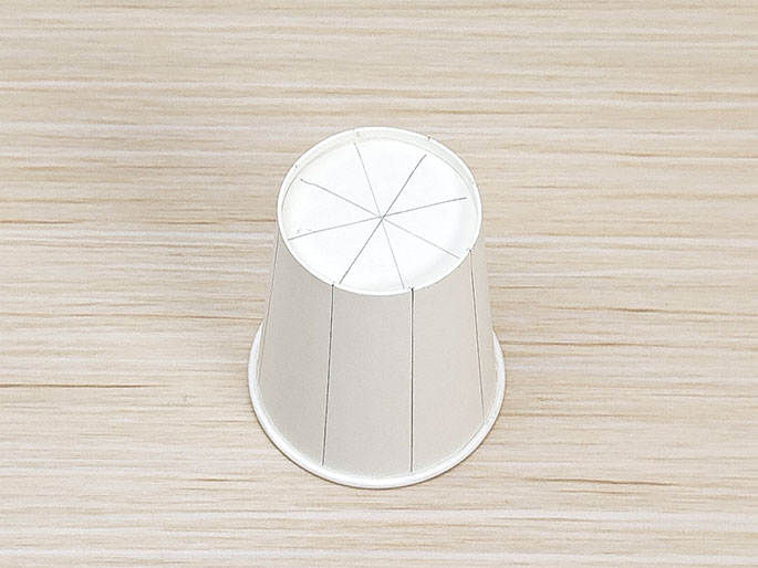 A small white paper cup is upside down on a light wooden surface. Lines are drawn on the bottom of the cup, dividing it into eight sections. Additional vertical lines are drawn on the sides of the cup, aligning with the lines on the bottom, indicating planned cuts or folds for a craft project.