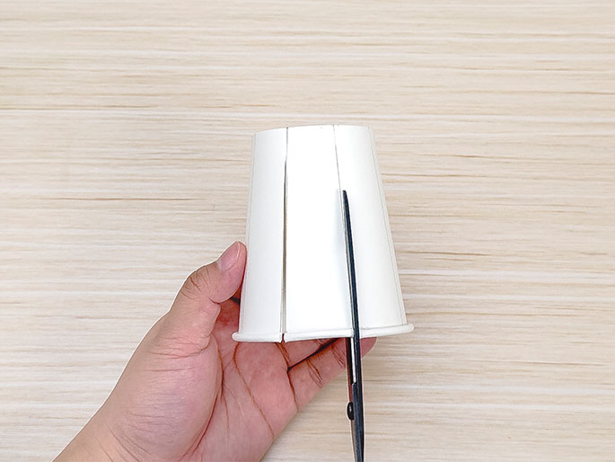 A hand holds a small white paper cup against a light wooden table. Scissors are cutting along a line drawn on the cup. The cup has several vertical lines marked on it, indicating it is being cut into sections as part of a craft project.