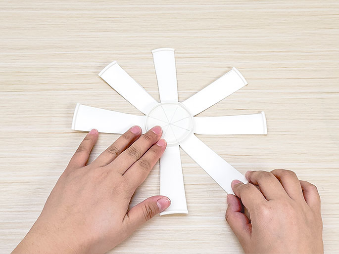 Two hands flatten a cut paper cup on a light wood surface. The cup has been cut into eight triangular segments, which are spread out like the spokes of a wheel, connected at the center circle of the cup's bottom. This shows a stage in progress of a paper craft project.