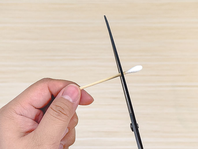A hand holds a cotton swab horizontally over a light wooden surface. Scissors are positioned to cut the cotton swab in half. The image focuses on the action of cutting the swab, likely for a craft or other project.