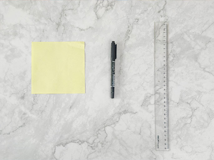 A blank pale yellow sticky note, a black fine-tipped marker, and a clear plastic ruler are arranged on a marbled gray surface. The items are laid out separately, suggesting preparation for a task involving measuring and marking, likely note-taking or a crafting project. The overhead view presents the items simply against the textured background.