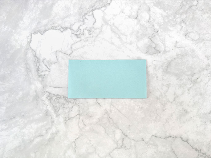 A rectangular piece of light teal or aqua-colored material lies flat on a marbled gray surface. The material appears to be paper or thin cardstock and is positioned horizontally. It is unmarked and takes up a small portion of the frame, with the marbled background providing a textured contrast. The image suggests the material is ready for use in a craft or other project.