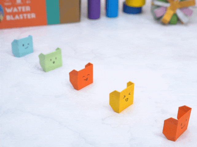 Four small origami boxes, each a different color (light green, orange, yellow, and orange-red), are arranged on a marble-like surface. Each box has a simple, hand-drawn face expressing a different emotion. Blurred in the background are various craft supplies, including a box labeled Water Blaster. The video showcases these small, folded paper creations.