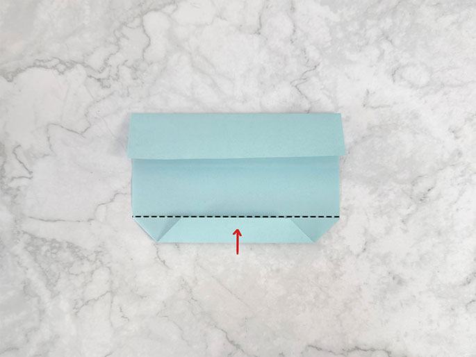 A light teal piece of paper or cardstock is being folded into a pouch or envelope shape on a marbled gray background. The top flap is folded down, and the bottom corners, previously folded inward, are now folded upwards, creating a trapezoidal shape at the bottom. A red arrow in the center indicates the upward folding motion of the bottom section. A dashed black line across this section likely marks a previous fold line or serves as a guide. The image illustrates a step in an origami or paper-folding process.