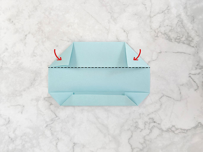 A light teal piece of paper is being folded on a marbled gray surface, resembling a flattened, open pouch or envelope. The top corners are folded downwards towards the center, and the bottom section is folded upwards. Red curved arrows on the top corners indicate the downward folding action. A dashed black line horizontally across the center likely indicates a previous fold line or serves as a guide for further folding. The image captures a stage in an origami or paper crafting process.