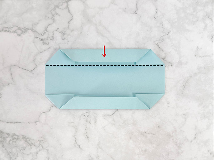 A light teal piece of paper or cardstock is folded on a marbled gray surface, resembling a narrow, open container or pouch. Both the top and bottom edges are folded inward and then folded down towards the center, creating small flaps. A red arrow pointing downwards indicates the folding direction of the top flap. Dashed black lines across the top and bottom near the folded edges likely indicate previous fold lines or guides for the current folds. The image shows a step-by-step process of paper folding, likely origami or a similar craft.