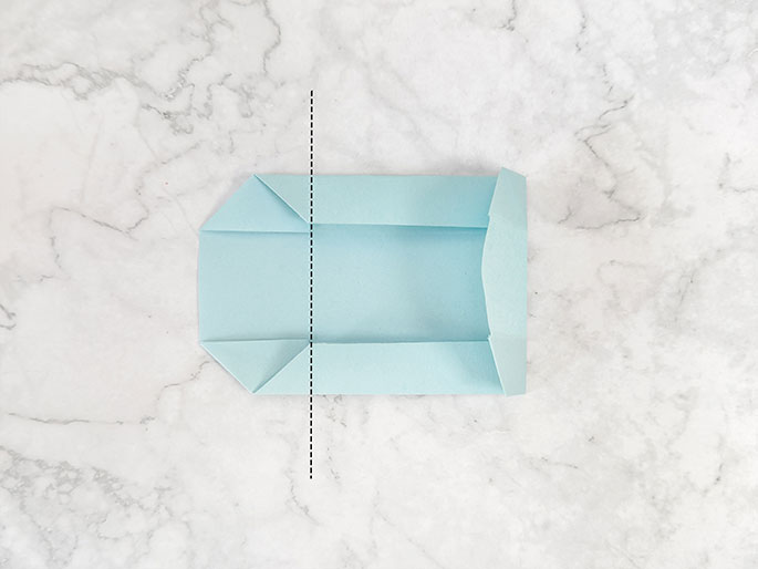 A light teal piece of paper, folded into a small, open container or pouch shape, rests on a marbled gray surface. The top and bottom edges are folded inwards and downwards towards the center, creating small flaps. A vertical dashed black line down the center of the paper suggests the next fold line for creating the final shape of the pouch or container. The image illustrates a step in a paper folding activity, likely origami or a similar craft.