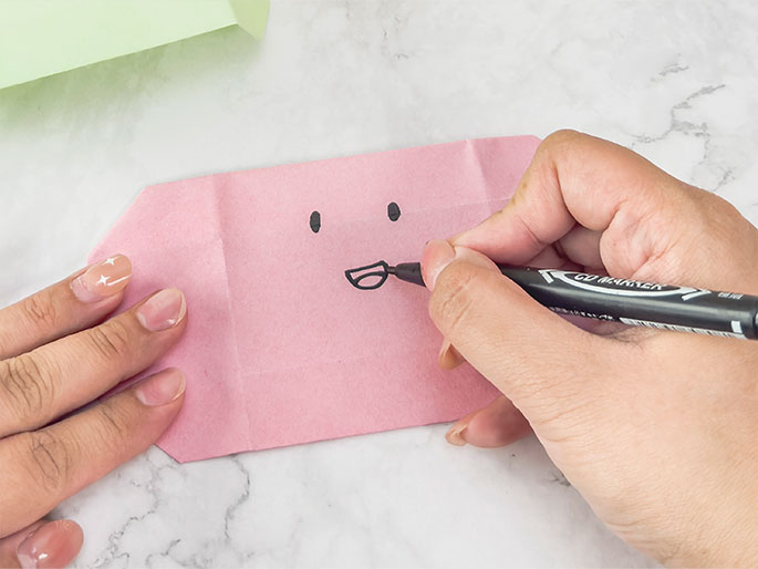 A person's hands are in the process of drawing a simple face onto a folded piece of light pink paper. The paper, possibly origami, resembles a small, flattened pouch. The face consists of two small oval eyes and a smiling mouth being drawn with a black marker. The hand holding the marker is visible in the right side of the frame, while the other hand gently holds the pink paper. A folded piece of light green paper is visible in the top left corner. The background is a marbled gray surface.