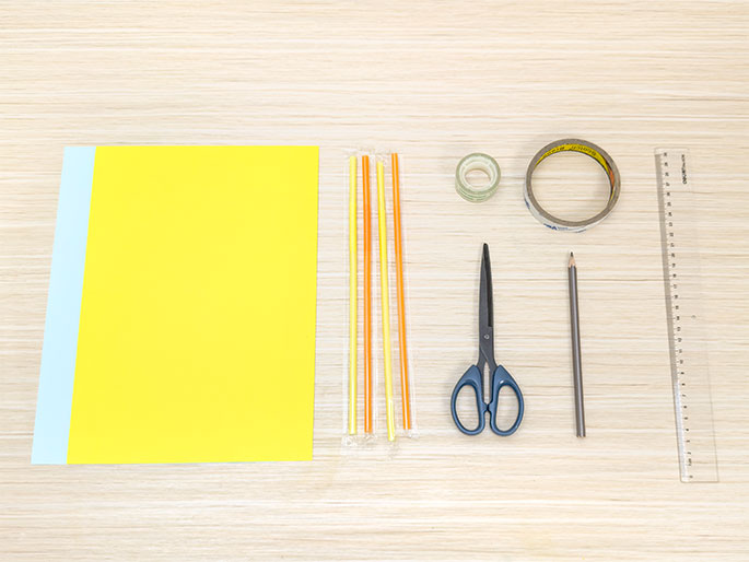 An assortment of craft supplies lies on a light woodgrain table, arranged for a project. The items include a large sheet of bright yellow construction paper, a group of four colorful drinking straws (orange and yellow), a roll of clear tape, a roll of double-sided tape, a pair of small blue scissors, a gray pencil, and a clear plastic ruler.