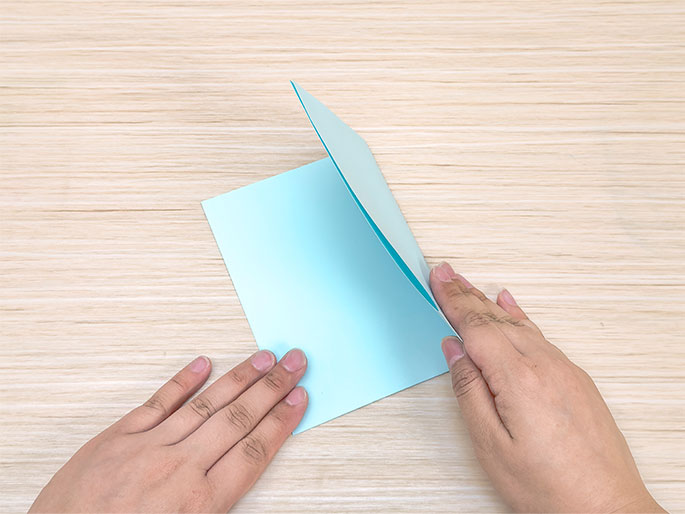 Two hands are folding a piece of light blue paper in half against a light woodgrain background. The paper is oriented in a landscape position, and the right side is being folded over to meet the left. The hands hold the paper near the top and bottom edges of the fold.