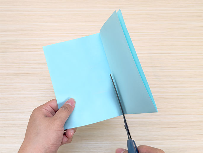 Two hands are shown cutting a folded piece of light blue paper with scissors against a light woodgrain background. The paper is folded in half, and the scissors are making a cut from the folded edge towards the open edges. The left hand holds the paper, and the right hand operates the scissors.