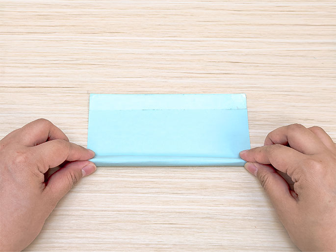 Two hands fold a rectangular piece of light blue paper in half lengthwise against a light woodgrain background. The top edge of the paper has a slightly darker blue band where it was previously folded. The hands hold the paper near the short ends, pinching the folded edge together.