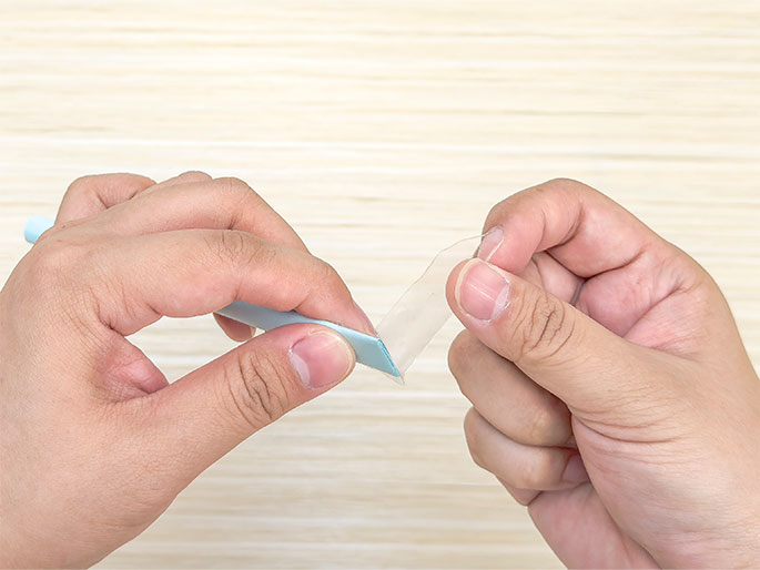 Two hands are shown applying a piece of clear tape to the end of a light blue folded piece of paper. The background is a light woodgrain surface. The left hand holds the paper, while the right hand holds the tape, which is partially attached to the paper's end. The tape is being peeled back from its roll.