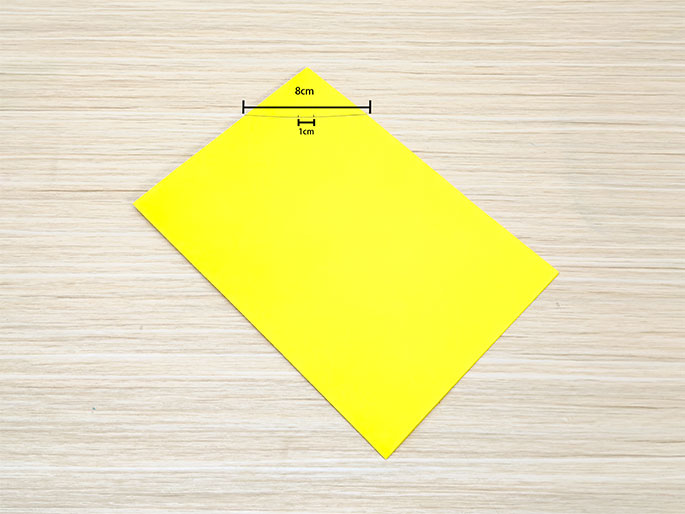 A bright yellow piece of rectangular paper rests on a light woodgrain table. The paper is positioned diagonally. Two lines with measurements indicate a section at the top edge of the paper. One line labeled 8cm spans a portion of the top edge, and a shorter perpendicular line labeled 1cm extends down from the midpoint of the 8cm line. This suggests markings for a cut or fold.