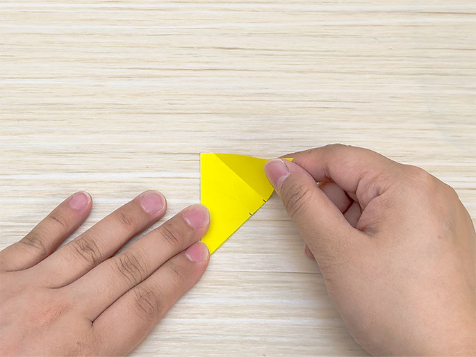 Two hands are folding a bright yellow piece of paper on a light woodgrain table. The paper is being folded into a triangular shape. The left hand holds the base of the triangle, while the right hand folds the top corner down. A dotted line is visible along the fold line.