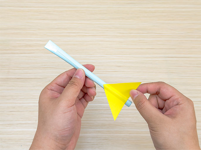 Two hands hold a light blue paper straw with a bright yellow paper triangle attached near one end. The straw appears to be made from rolled paper, and a small piece of white tape is visible near the open end. The yellow triangle is positioned to resemble the fins of a rocket or dart. The hands are manipulating the yellow triangle, seemingly attaching or adjusting it onto the straw. The background is a light woodgrain surface.
