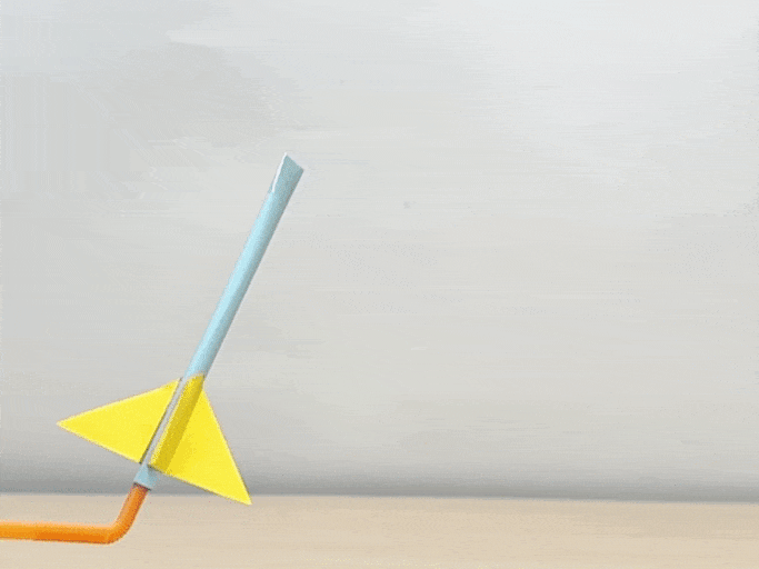A simple straw rocket is launched using another straw. The rocket consists of a light blue drinking straw with a yellow triangular fin attached. An orange straw is used to blow air into the rocket, propelling it upwards against a white background. The rocket spins and tumbles as it flies through the air. A second rocket, identical in construction but with a red fin, is subsequently launched in the same manner. A popping sound is heard as the rockets are launched.