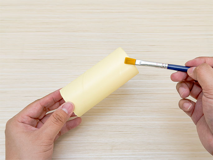 Two hands are shown above a light wooden surface. One hand holds a pale yellow cardboard tube, while the other hand holds a paintbrush with a blue handle and applies yellow paint to the tube. The focus is on the process of painting the cardboard tube.