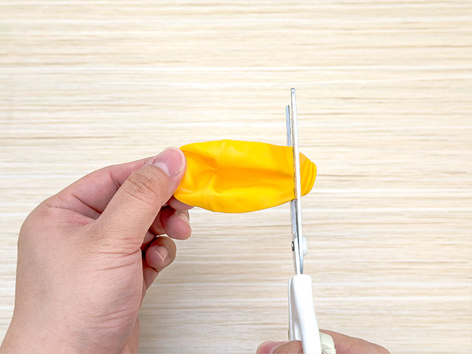 Two hands are shown cutting a deflated yellow balloon with scissors against a light wooden background. The balloon is being held taut, and the scissors are positioned to cut off the end opposite the opening. The focus is on the action of cutting the balloon as part of a craft or project.