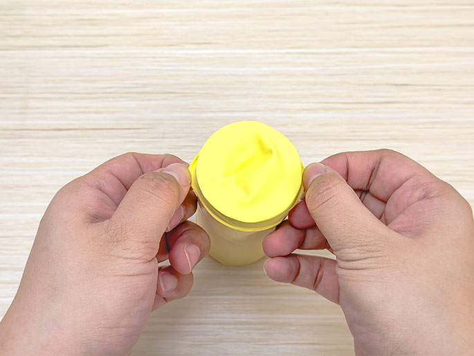 Two hands are stretching a piece of yellow balloon material over one end of a cardboard tube against a light wooden background. It appears the balloon material is being used to create a drum-like membrane or covering for the tube.